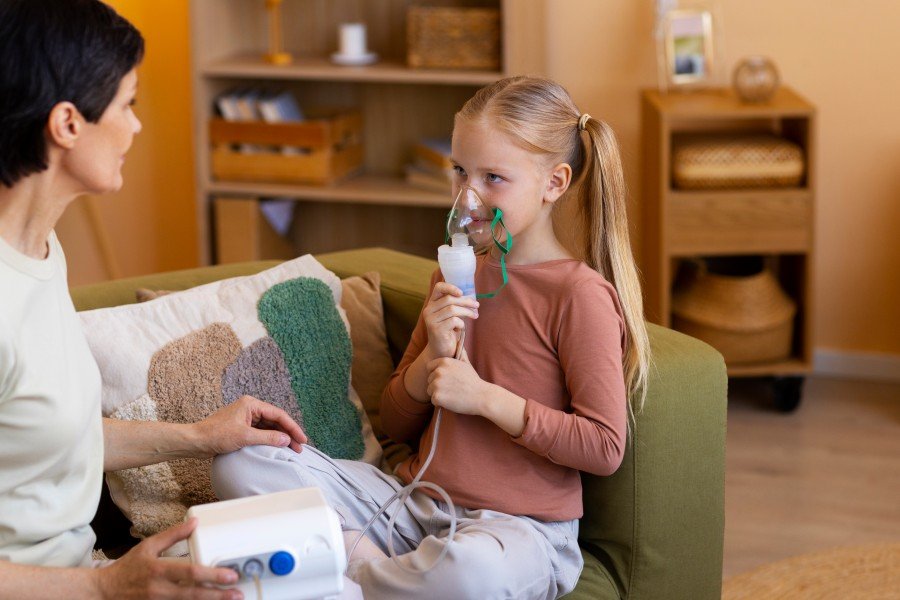 Young girl using a nebulizer for asthma treatment while sitting with her mother at home.