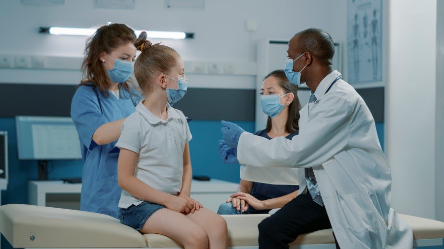 A diverse medical team in a clinic examines a young girl while a masked doctor gestures toward an unseen medical image.