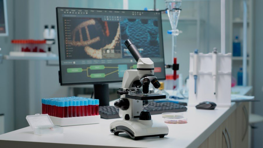 Laboratory microscope on a workbench surrounded by blood sample tubes, lab equipment, and a computer displaying scientific images